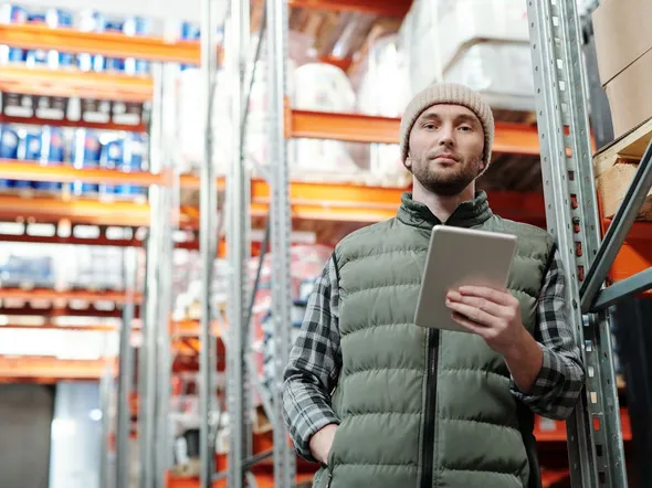 Young male warehouse manager in a beanie holding a tablet in a storeroom.
