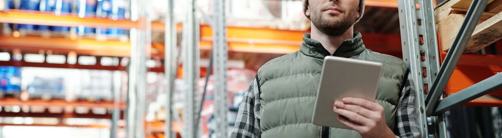 Young male warehouse manager in a beanie holding a tablet in a storeroom.