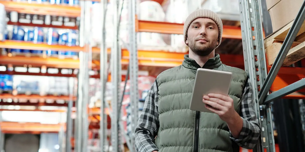 Young male warehouse manager in a beanie holding a tablet in a storeroom.