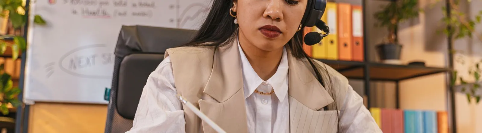 Professional woman wearing a headset taking notes in an office setting, focused on work.