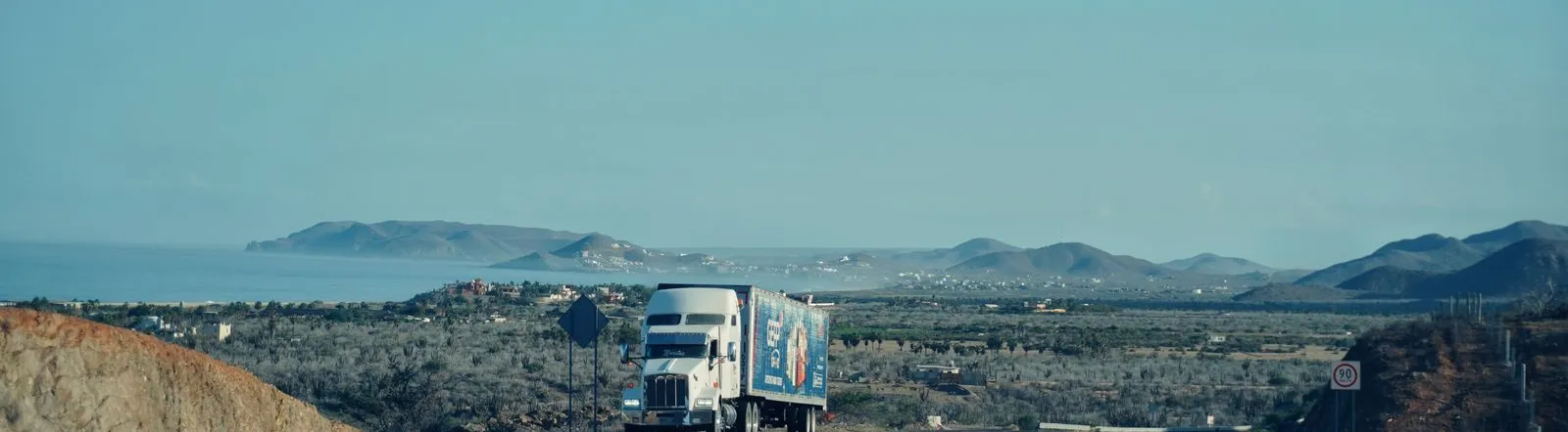 A semi-truck travels on a scenic highway with mountains in the background under a clear sky.