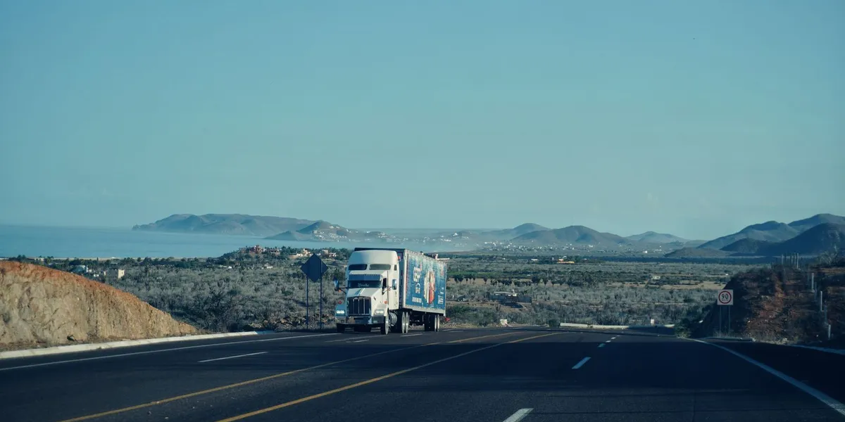 A semi-truck travels on a scenic highway with mountains in the background under a clear sky.
