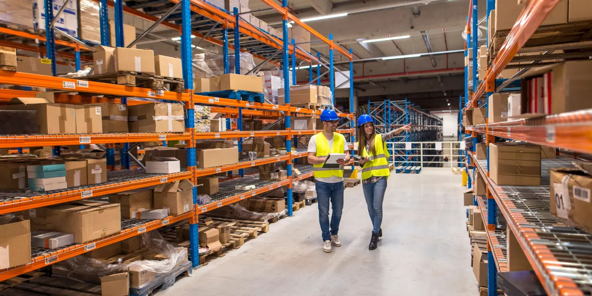 Two workers in safety vests walking through a warehouse aisle checking inventory