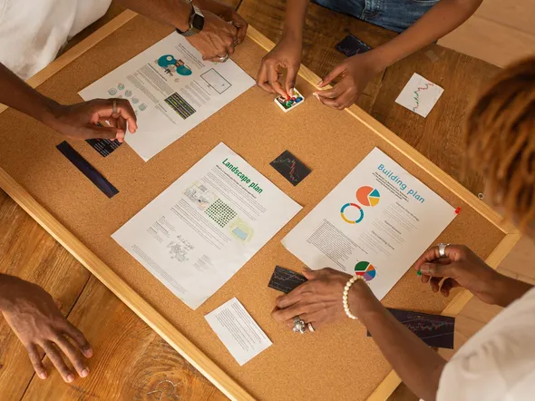 A diverse team collaborates on a workspace board with charts and plans.